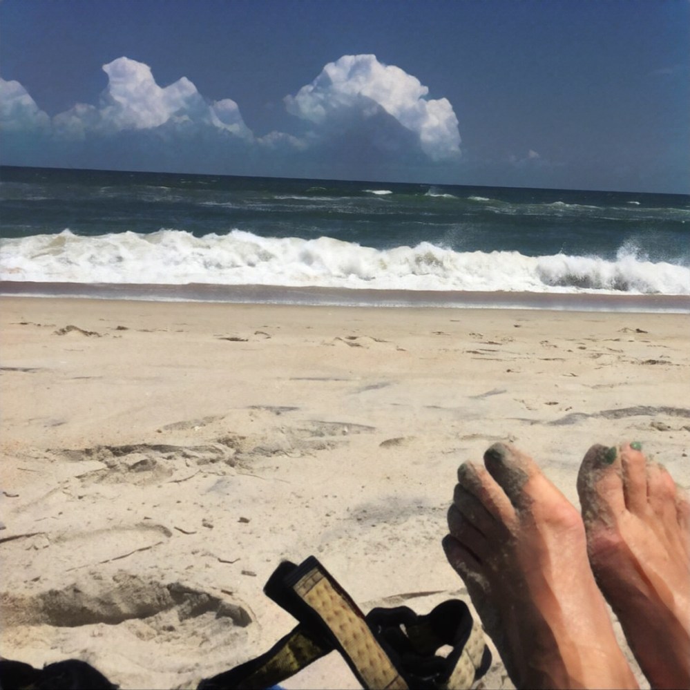 The waves are crashing on the beach. There are some big fluffy clouds. And some sandy feet and sandy sandals in the foreground. It's sunny, too.