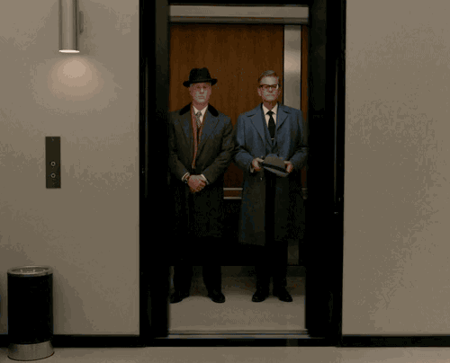 A nondescript elevator with two white guys from the 60s madmen era with fedoras with the doors closing.
