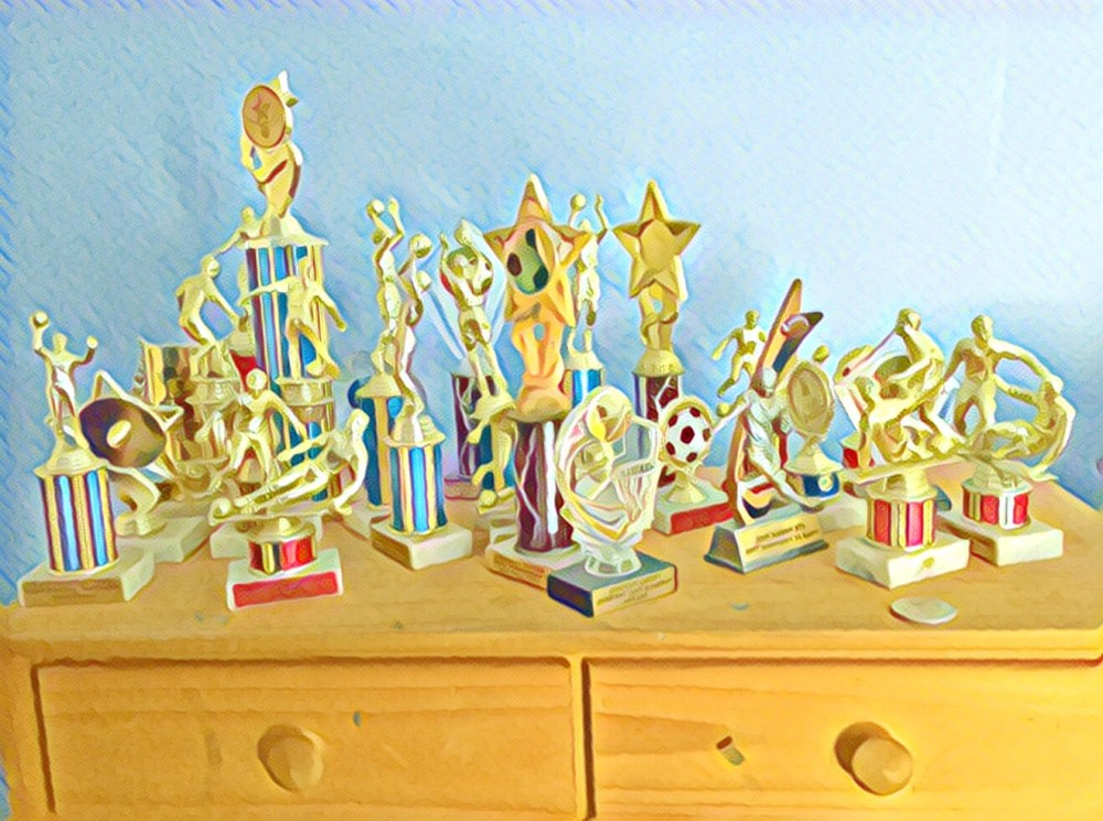 A dresser covered with two dozen trophies from basketball, soccer, rugby, baseball and football. Some personalized, most generic. 